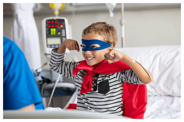 A little boy sitting in a hospital bed wearing a superhero cape and holding his arms up in the air.